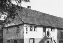 FELDER DER IDENTITÄT – SPAZI DI IDENTITÀ II / Kulturforum Rom Das SW-Bild zeigt ein älteres, langgestrecktes Wohnhaus mit einem steilen Treppenaufgang, auf dem eine Frau steht. Sie trägt eine Schürze und einen Haarknoten. Aus einem der Fenster schaut ein älterer Mann.