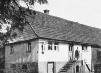 FELDER DER IDENTITÄT – SPAZI DI IDENTITÀ II / Kulturforum Rom Das SW-Bild zeigt ein älteres, langgestrecktes Wohnhaus mit einem steilen Treppenaufgang, auf dem eine Frau steht. Sie trägt eine Schürze und einen Haarknoten. Aus einem der Fenster schaut ein älterer Mann.