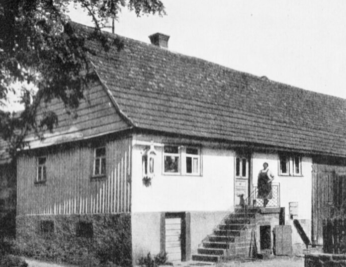 Bild: Gaia Credentino Das SW-Bild zeigt ein älteres, langgestrecktes Wohnhaus mit einem steilen Treppenaufgang, auf dem eine Frau steht. Sie trägt eine Schürze und einen Haarknoten. Aus einem der Fenster schaut ein älterer Mann.