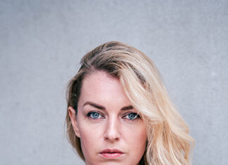 Valerie Fritsch / Botschaft Oslo Das Bild zeigt die Autorin Valerie Fritsch vor grauem Hintergrund. Sie trägt eine schwarzen Bluse mit V-Ausschnitt und schaut gerade in die Kamera. Sie hat blaue Augen und ihr schulterlanges blondes Haar trägt sie mit Seitenscheitel.