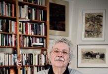 Karl-Markus Gauß / Abenteurer des Schreibens Auf dem Bild sieht man Karl Markus Gauss stehend vor einem Bücherschrank. Er trägt einen grauen Pullover und schwarze Hose und hat die Hände vor der Brust überkreuzt. Er hat weisse lockige Haare und einen Schnauzbart und trägt eine Brille.