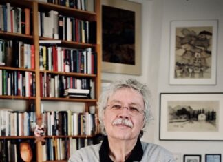 Karl-Markus Gauß / Abenteurer des Schreibens Auf dem Bild sieht man Karl Markus Gauss stehend vor einem Bücherschrank. Er trägt einen grauen Pullover und schwarze Hose und hat die Hände vor der Brust überkreuzt. Er hat weisse lockige Haare und einen Schnauzbart und trägt eine Brille.