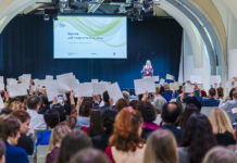 Culture as a peace act / Gemeinsame Erklärung Das Bild zeigt Inna Shevchenko auf der Bühne. Neben ihr der Screen mit der Ankündigung der Keynote. Vor ihr das Publikum hält weisse Blätter in die Höhe.