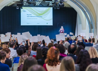 Culture as a peace act / Gemeinsame Erklärung Das Bild zeigt Inna Shevchenko auf der Bühne. Neben ihr der Screen mit der Ankündigung der Keynote. Vor ihr das Publikum hält weisse Blätter in die Höhe.