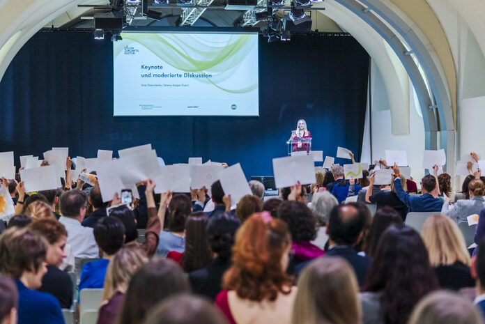 Das Bild zeigt Inna Shevchenko auf der Bühne. Neben ihr der Screen mit der Ankündigung der Keynote. Vor ihr das Publikum hält weisse Blätter in die Höhe.