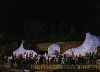 Multimediale Performance / Kulturforum Sarajewo Das Bild zeigt eine beleuchtete Wand mit stehenden Menschen vor dunklem Nachthimmel.