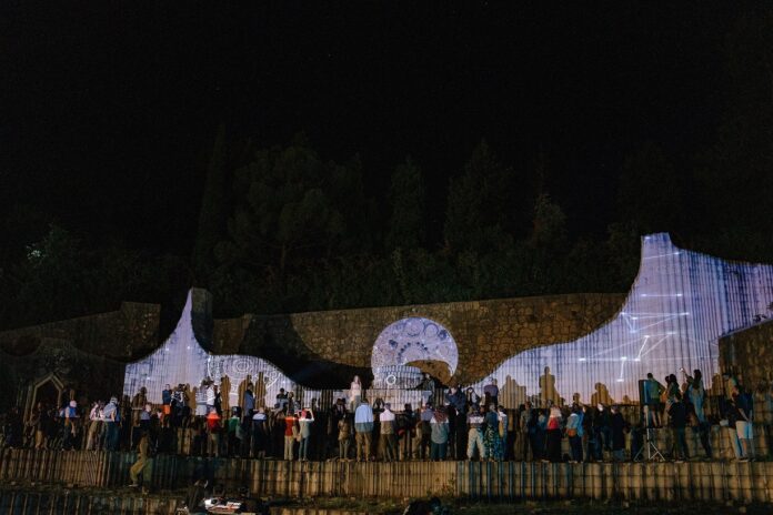 Das Bild zeigt eine beleuchtete Wand mit stehenden Menschen vor dunklem Nachthimmel.