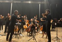 No Borders Orchestra / Botschaft Pristina Das Bild zeigt Dirigent und Musiker des Orchesters in einer Konzerthalle.