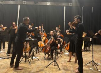 No Borders Orchestra / Botschaft Pristina Das Bild zeigt Dirigent und Musiker des Orchesters in einer Konzerthalle.