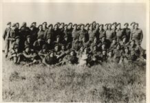 Austrian Refugees in the British Army /Kulturforum London Das Bild zeigt die No.3 Troop im Mai 1943. Es ist ein leicht vergilbtes schwarz-weiß Foto von Soldaten in Uniform, im Vordergrund ist hohes Gras. In der Mitte vorne ist auch ein weißer Hund zu erkennen. Viele Soldaten lächeln in die Kamera und wirken entspannt.