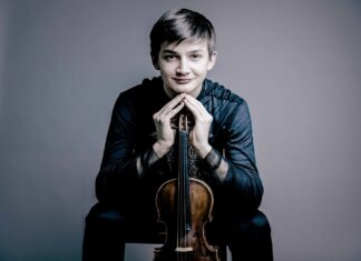 Feierliches Konzert / Kulturforum Zagreb Das Bild zeigt den jungen Geiger Leonhard Baumgartner. In seinen Händen hält er eine Violine, die senkrecht vor ihm steht, mit dem Griffbrett nach oben. Seine Hände sind an den Fingerspitzen zusammengelegt und ruhen auf dem oberen Ende der Violine.