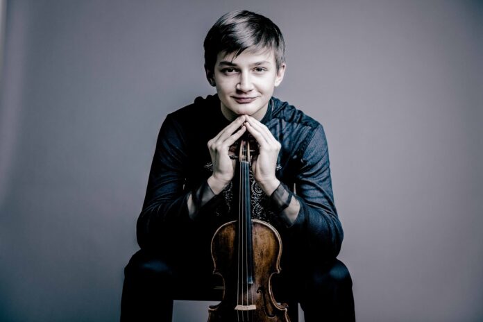 Bild: @ Julia Wesely / Andrej Grilc / NRK Das Bild zeigt den jungen Geiger Leonhard Baumgartner. In seinen Händen hält er eine Violine, die senkrecht vor ihm steht, mit dem Griffbrett nach oben. Seine Hände sind an den Fingerspitzen zusammengelegt und ruhen auf dem oberen Ende der Violine.