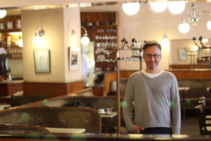 Eugene Quinn Auf dem Foto ist Eugene Quinn zu sehen. Er steht in einem traditionellen Wiener Kaffeehaus – einem Ort, der auch in seinem Projekt Coffeehouse Conversations eine zentrale Rolle spielt.