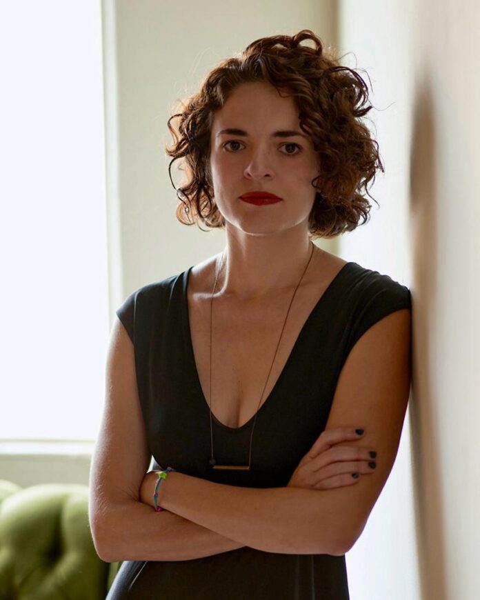 Das Bild zeigt die Kuratorin Lorena Moreno Vera. Sie hat kurze wellige dunkle Haare und trägt ein ärmelloses dunkles Kleid. Sie lehnt an einer Wand, hat die Hände vorne überkreuzt und blickt direkt in die Kamera.