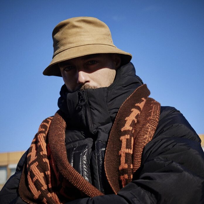 © Denis Laner Auf dem Bild ist Andi Stecher zu sehen. Er trägt einen braunen Schlapphut, der einen Schatten über sein Gesicht wirft. Einen dicken schwarzen Anorak und einen braunen Schal.