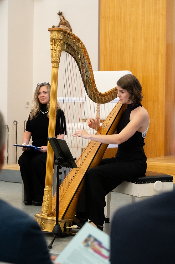 Auf dem Bild ist die Musikerin auf der Harfe spielend zu sehen. Dahinter sitzt Beata Greneche mit einem Buch vor ihr.