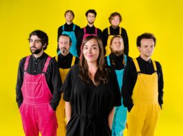 Yasmo & die Klangkantine / Kulturforum Berlin Auf dem Foto steht eine Frau im Vordergrund und lächelt leicht in die Kamera. Sie trägt ein schwarzes Kleid und hat lange, braune Haare. Hinter ihr stehen sieben Männer in zwei Reihen. Alle tragen schwarze Hemden und farbige Latzhosen in Pink, Gelb oder Türkis. Die Personen blicken überwiegend neutral oder ernst nach vorne. Der Hintergrund ist ein gleichmäßig leuchtendes Gelb, das dem Bild einen sehr farbintensiven, kontrastreichen Gesamteindruck verleiht.