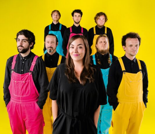 Yasmo & die Klangkantine / Kulturforum Berlin Auf dem Foto steht eine Frau im Vordergrund und lächelt leicht in die Kamera. Sie trägt ein schwarzes Kleid und hat lange, braune Haare. Hinter ihr stehen sieben Männer in zwei Reihen. Alle tragen schwarze Hemden und farbige Latzhosen in Pink, Gelb oder Türkis. Die Personen blicken überwiegend neutral oder ernst nach vorne. Der Hintergrund ist ein gleichmäßig leuchtendes Gelb, das dem Bild einen sehr farbintensiven, kontrastreichen Gesamteindruck verleiht.