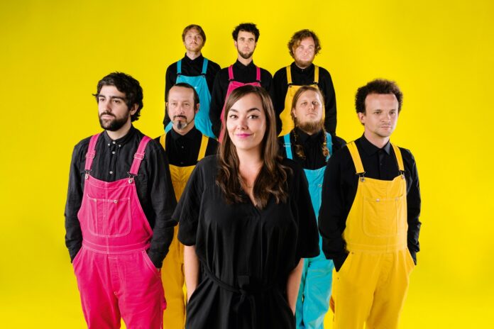 Bild: © Karo Pernegger Auf dem Foto steht eine Frau im Vordergrund und lächelt leicht in die Kamera. Sie trägt ein schwarzes Kleid und hat lange, braune Haare. Hinter ihr stehen sieben Männer in zwei Reihen. Alle tragen schwarze Hemden und farbige Latzhosen in Pink, Gelb oder Türkis. Die Personen blicken überwiegend neutral oder ernst nach vorne. Der Hintergrund ist ein gleichmäßig leuchtendes Gelb, das dem Bild einen sehr farbintensiven, kontrastreichen Gesamteindruck verleiht.