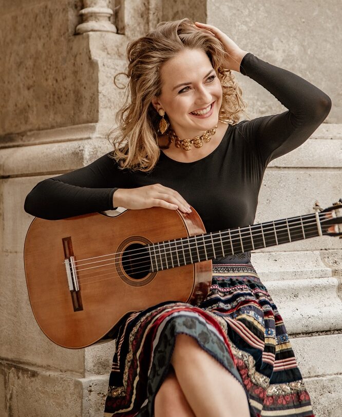 Bild: Horacio Reyes Paéz Das Bild zeigt die österreichische Gitarristin Rosa Franziska Maier mit der Gitarre in der Hand. Sie hat langes blondes Haar, trägt ein grünes Shirt und einen bunten Rock. Mit einer Hand streicht sie sich die Haare zurück, mit der anderen hält sie die Gitarre auf ihrem Oberschenkel, sie lacht.