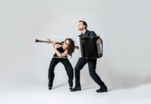 Duo Minerva: Hymne à l‘amour / Kulturforum New York Zwei Musiker*innen stehen vor einem hellgrauen Hintergrund. Links ist eine Person mit lockigem, schulterlangem Haar zu sehen, die eine Klarinette spielt. Sie trägt schwarze Kleidung und beugt sich dynamisch nach vorne. Hinter ihr hält eine zweite Person, ebenfalls in schwarzer Kleidung, ein großes Akkordeon und spielt es. Beide wirken in Bewegung, als würden sie eine energiegeladene musikalische Performance darstellen.