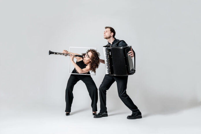 Zwei Musiker*innen stehen vor einem hellgrauen Hintergrund. Links ist eine Person mit lockigem, schulterlangem Haar zu sehen, die eine Klarinette spielt. Sie trägt schwarze Kleidung und beugt sich dynamisch nach vorne. Hinter ihr hält eine zweite Person, ebenfalls in schwarzer Kleidung, ein großes Akkordeon und spielt es. Beide wirken in Bewegung, als würden sie eine energiegeladene musikalische Performance darstellen.