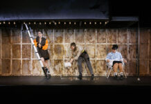 Verbranntes Land / Kulturforum Prag Auf dem Foto ist eine Bühne zu sehen, die von einer Reihe an der Decke befestigten nackten Glühbirnen beleuchtet wird. Auf der Bühne befinden sich drei junge Frauen in trendiger Alltagskleidung Die linke lehnt an einer Leiter, die mittlere gießt gerade Wasser aus einem weißen Plastikeimer auf den Boden der Bühne, und die rechte sitzt in einem weißen Korbstuhl und beobachtet das vergossene Wasser.