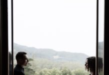 Konzertreise des Klavierduos Kalabova-Gugg / Kulturforum Prag Das Foto zeigt die Umrisse eines Mannes und einer Frau, die jeweils links und rechts von einer offenen Balkontür stehen. Zwischen ihnen ist Balkongeländer sowie Ausblick auf eine Lichtung und einen Hügel voller Bäume in hellem Licht zu sehen.