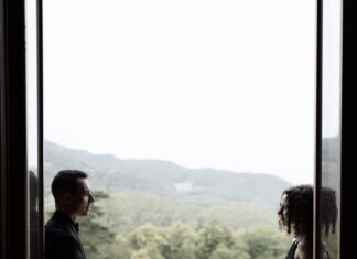 Konzertreise des Klavierduos Kalabova-Gugg / Kulturforum Prag Das Foto zeigt die Umrisse eines Mannes und einer Frau, die jeweils links und rechts von einer offenen Balkontür stehen. Zwischen ihnen ist Balkongeländer sowie Ausblick auf eine Lichtung und einen Hügel voller Bäume in hellem Licht zu sehen.