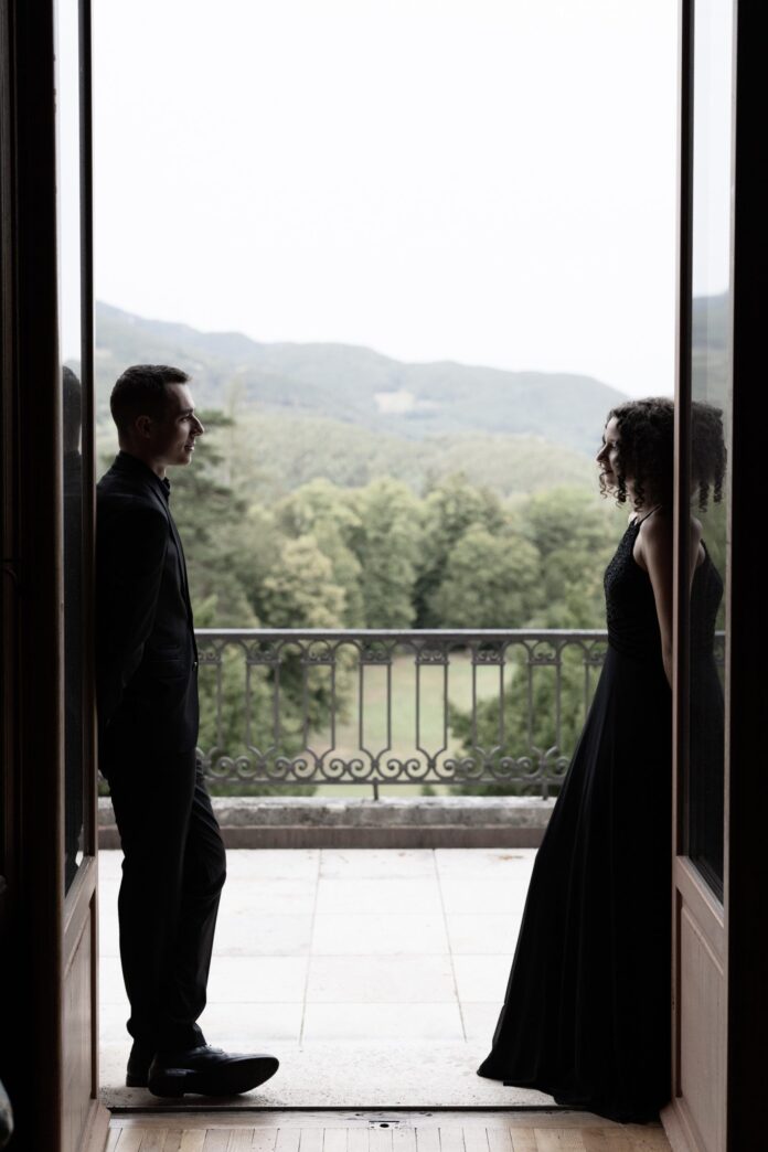 Das Foto zeigt die Umrisse eines Mannes und einer Frau, die jeweils links und rechts von einer offenen Balkontür stehen. Zwischen ihnen ist Balkongeländer sowie Ausblick auf eine Lichtung und einen Hügel voller Bäume in hellem Licht zu sehen.