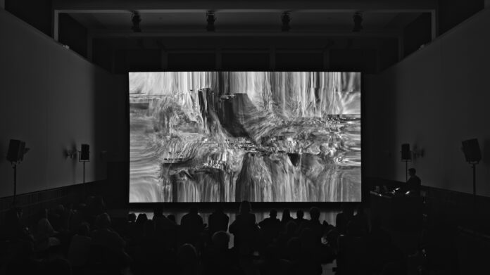 Auf dem Bild ist in Dunkel die Bühne zu sehen, darauf eine große Leinwand, auf der in SchwarzWEiss ein verschwommenes Bild zu sehen ist. Der Künstler steht auf der Seite mit einem Keyboard.