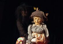Crazy Old Me / Kulturforum Istanbul Das Bild zeigt eine Puppentheaterszene: Eine Person in Schwarz führt eine alte Frau als Puppe mit Brille und Haube. Davor steht ein Plattenspieler mit einem bunt bemalten Totenschädel. Der Hintergrund ist dunkel, die Figuren sind warm beleuchtet.