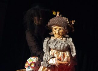 Crazy Old Me / Kulturforum Istanbul Das Bild zeigt eine Puppentheaterszene: Eine Person in Schwarz führt eine alte Frau als Puppe mit Brille und Haube. Davor steht ein Plattenspieler mit einem bunt bemalten Totenschädel. Der Hintergrund ist dunkel, die Figuren sind warm beleuchtet.