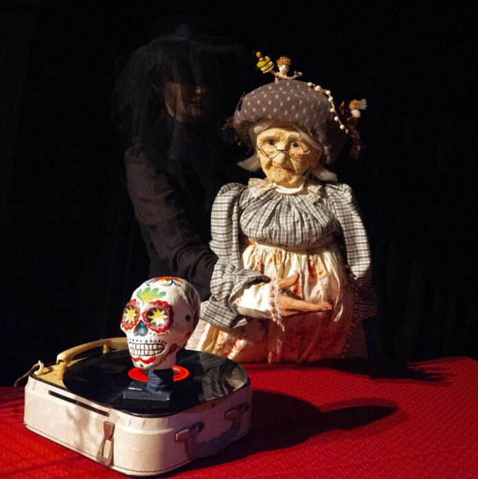 Das Bild zeigt eine Puppentheaterszene: Eine Person in Schwarz führt eine alte Frau als Puppe mit Brille und Haube. Davor steht ein Plattenspieler mit einem bunt bemalten Totenschädel. Der Hintergrund ist dunkel, die Figuren sind warm beleuchtet.
