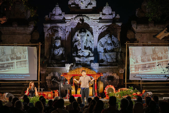 Das Bild zeigt die Szene des Stücks in Ubud aus der Zuseherperspektive vor einer beeindruckenden javanischen Tempelkulisse. Im Zentrum ist der Schauspieler Marten Schmidt als Erzähler, links die Musikerin Anna Anderluh, rechts der Musiker Xinwei Thow und dahinter das Gamelan Orchester.