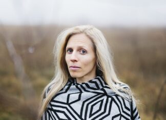 Sophie Reyer: Die Vielschreiberin / Podcast wissensArt Das Bild zeigt ein Portrait von Sophie Reyer. Sie steht in einer Landschaft, die verschwommen im Hintergrund sichtbar ist. Sie hat lange blonde Haare und blickt aufmerksam in die Kamera.