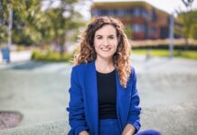 Das Fluchtparadox / Kulturforum Paris Das Bild zeigt Judith Kohlenberger. Sie trägt einen blitzblauen Anzug und ein schwarzes Shirt. Ihre Haare sind brünett, lang und lockig. Sie sitze auf einer Mauer und lächelt freundlich in die Kamera.