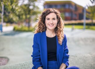 Das Fluchtparadox / Kulturforum Paris Das Bild zeigt Judith Kohlenberger. Sie trägt einen blitzblauen Anzug und ein schwarzes Shirt. Ihre Haare sind brünett, lang und lockig. Sie sitze auf einer Mauer und lächelt freundlich in die Kamera.