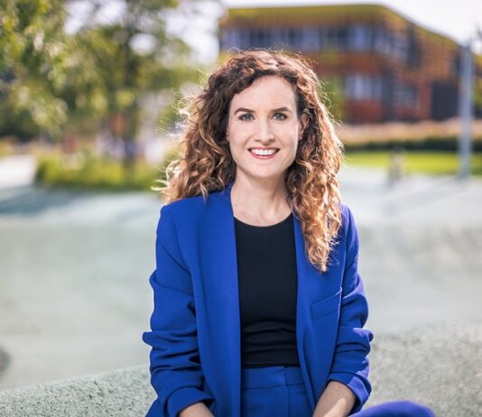 Das Fluchtparadox / Kulturforum Paris Das Bild zeigt Judith Kohlenberger. Sie trägt einen blitzblauen Anzug und ein schwarzes Shirt. Ihre Haare sind brünett, lang und lockig. Sie sitze auf einer Mauer und lächelt freundlich in die Kamera.