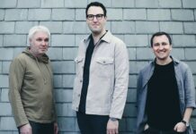 Owls – Konzertreise Polen / Kulturforum Warschau Das Bild zeigt die drei Mitglieder des österreichischen Jazztrios Owls vor einer grauen Schieferwand. Links steht ein Mann mit kurz geschnittenem, weißem Haar und einem beigefarbenen Hoodie. In der Mitte steht ein Mann mit dunklen Haaren und Brille, der eine helle, beige Jacke über dunkler Kleidung trägt. Rechts steht ein lächelnder Mann mit kurzen, dunklen Haaren in einem grauen Hemd und schwarzem T-Shirt. Alle drei blicken freundlich in die Kamera.