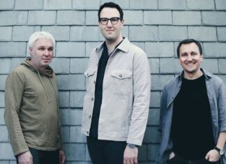 Owls – Konzertreise Polen / Kulturforum Warschau Das Bild zeigt die drei Mitglieder des österreichischen Jazztrios Owls vor einer grauen Schieferwand. Links steht ein Mann mit kurz geschnittenem, weißem Haar und einem beigefarbenen Hoodie. In der Mitte steht ein Mann mit dunklen Haaren und Brille, der eine helle, beige Jacke über dunkler Kleidung trägt. Rechts steht ein lächelnder Mann mit kurzen, dunklen Haaren in einem grauen Hemd und schwarzem T-Shirt. Alle drei blicken freundlich in die Kamera.