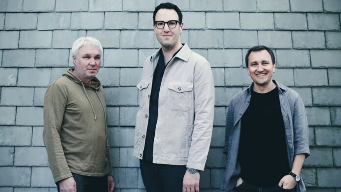 Das Bild zeigt die drei Mitglieder des österreichischen Jazztrios Owls vor einer grauen Schieferwand. Links steht ein Mann mit kurz geschnittenem, weißem Haar und einem beigefarbenen Hoodie. In der Mitte steht ein Mann mit dunklen Haaren und Brille, der eine helle, beige Jacke über dunkler Kleidung trägt. Rechts steht ein lächelnder Mann mit kurzen, dunklen Haaren in einem grauen Hemd und schwarzem T-Shirt. Alle drei blicken freundlich in die Kamera.