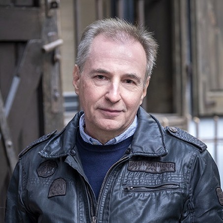 Das Bild zeigt ein Portrait von Alois Hotschnig. Er trägt eine schwarze Lederjacke, darunter einen Wollpullover und ein Hemd, seine Haare sind grau meliert, er lächelt in die Kamera.