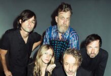 Deutschland-Tour: Naked Lunch / Kulturforum Berlin Das Bild zeigt ein Studiofoto der fünfköpfigen Band Naked Lunch vor grauem Hintergrund. In der ersten Reihe sitzen die Bassistin Romy Jakovcic sowie der Frontman Oliver Welter. Der Rest der Band ist stehend in der zweiten Reihe zu sehen (Wolfgang Lehmann, Boris Hauf und Alex Jezdinsky). Alle tragen überwiegend dunkle, elegante Kleidung und blicken ernst oder neutral in die Kamera. Die Aufnahme wirkt professionell und stilisiert.