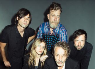 Deutschland-Tour: Naked Lunch / Kulturforum Berlin Das Bild zeigt ein Studiofoto der fünfköpfigen Band Naked Lunch vor grauem Hintergrund. In der ersten Reihe sitzen die Bassistin Romy Jakovcic sowie der Frontman Oliver Welter. Der Rest der Band ist stehend in der zweiten Reihe zu sehen (Wolfgang Lehmann, Boris Hauf und Alex Jezdinsky). Alle tragen überwiegend dunkle, elegante Kleidung und blicken ernst oder neutral in die Kamera. Die Aufnahme wirkt professionell und stilisiert.