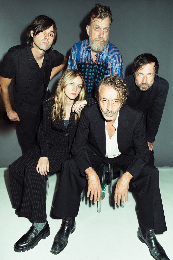 KF Berlin Das Bild zeigt ein Studiofoto der fünfköpfigen Band Naked Lunch vor grauem Hintergrund. In der ersten Reihe sitzen die Bassistin Romy Jakovcic sowie der Frontman Oliver Welter. Der Rest der Band ist stehend in der zweiten Reihe zu sehen (Wolfgang Lehmann, Boris Hauf und Alex Jezdinsky). Alle tragen überwiegend dunkle, elegante Kleidung und blicken ernst oder neutral in die Kamera. Die Aufnahme wirkt professionell und stilisiert.