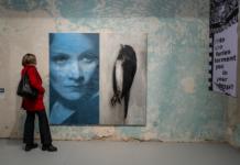 Why ‚We Should All Be Feminists’ / Kulturforum Bukarest Auf dem Bild sieht man eine Dame in einer roten Jacke die vor einem großen Gemälde in der Galerie steht. Das Gemälde zeigt ein Frauengesicht links, und einen Vogel rechts.