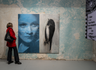 Why ‚We Should All Be Feminists’ / Kulturforum Bukarest Auf dem Bild sieht man eine Dame in einer roten Jacke die vor einem großen Gemälde in der Galerie steht. Das Gemälde zeigt ein Frauengesicht links, und einen Vogel rechts.