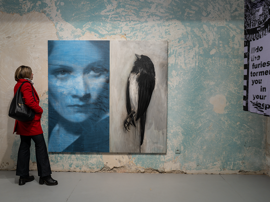 Why ‚We Should All Be Feminists’ / Kulturforum Bukarest Auf dem Bild sieht man eine Dame in einer roten Jacke die vor einem großen Gemälde in der Galerie steht. Das Gemälde zeigt ein Frauengesicht links, und einen Vogel rechts.