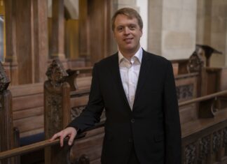 Orgelklänge am Bosporus / Kulturforum Istanbul Das Bild zeigt Stefan Donner in dunklem Anzug, der lächelnd in einem Kirchenraum mit hölzernen Sitzbänken steht und in die Kamera blickt.